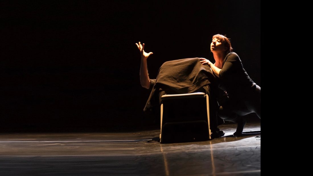 Sur une scène sombre une femme semble danser près d'une chaise où un homme se ca