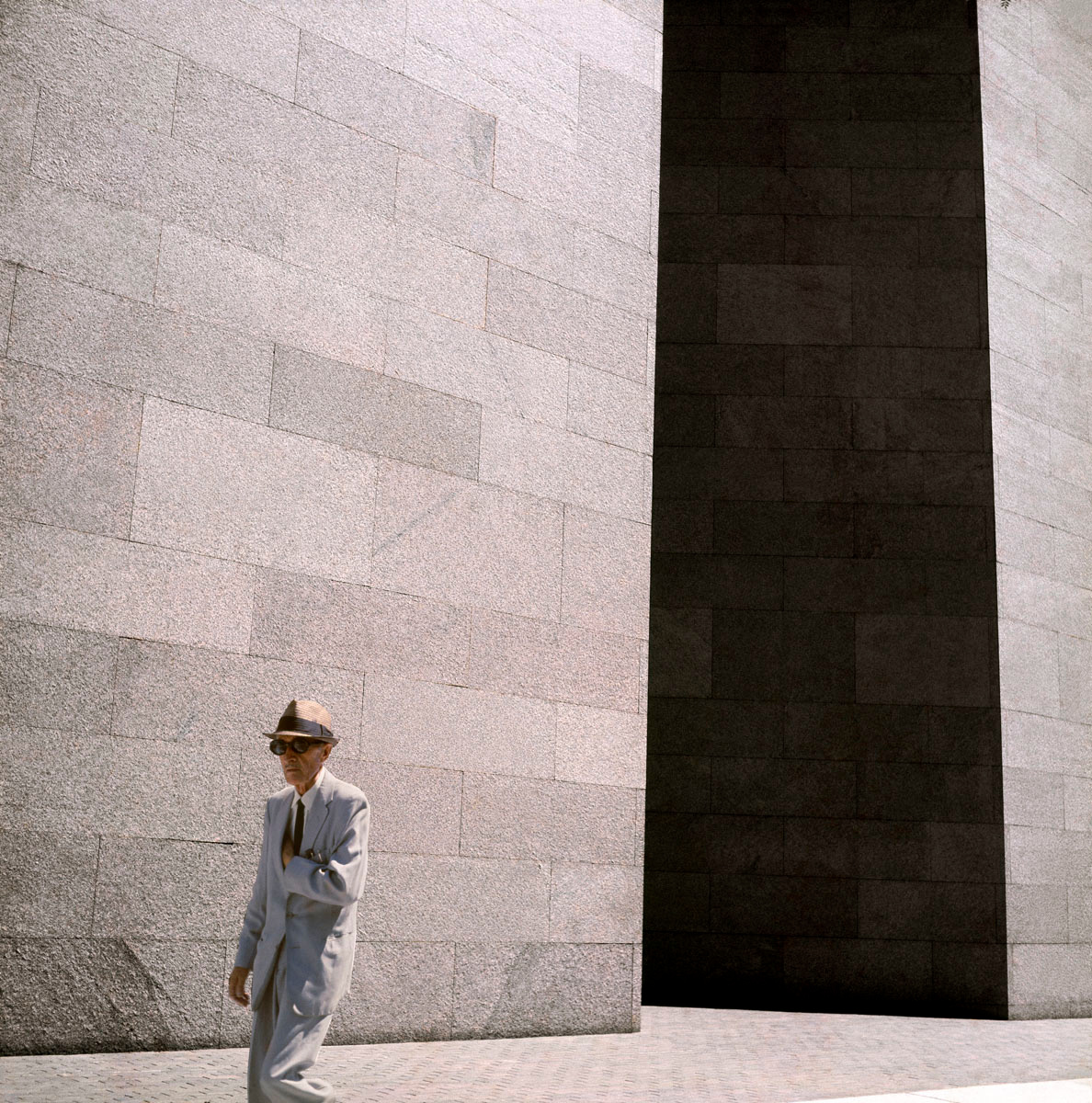 La photographie montre un homme chapeauté marchant devant un immeuble.