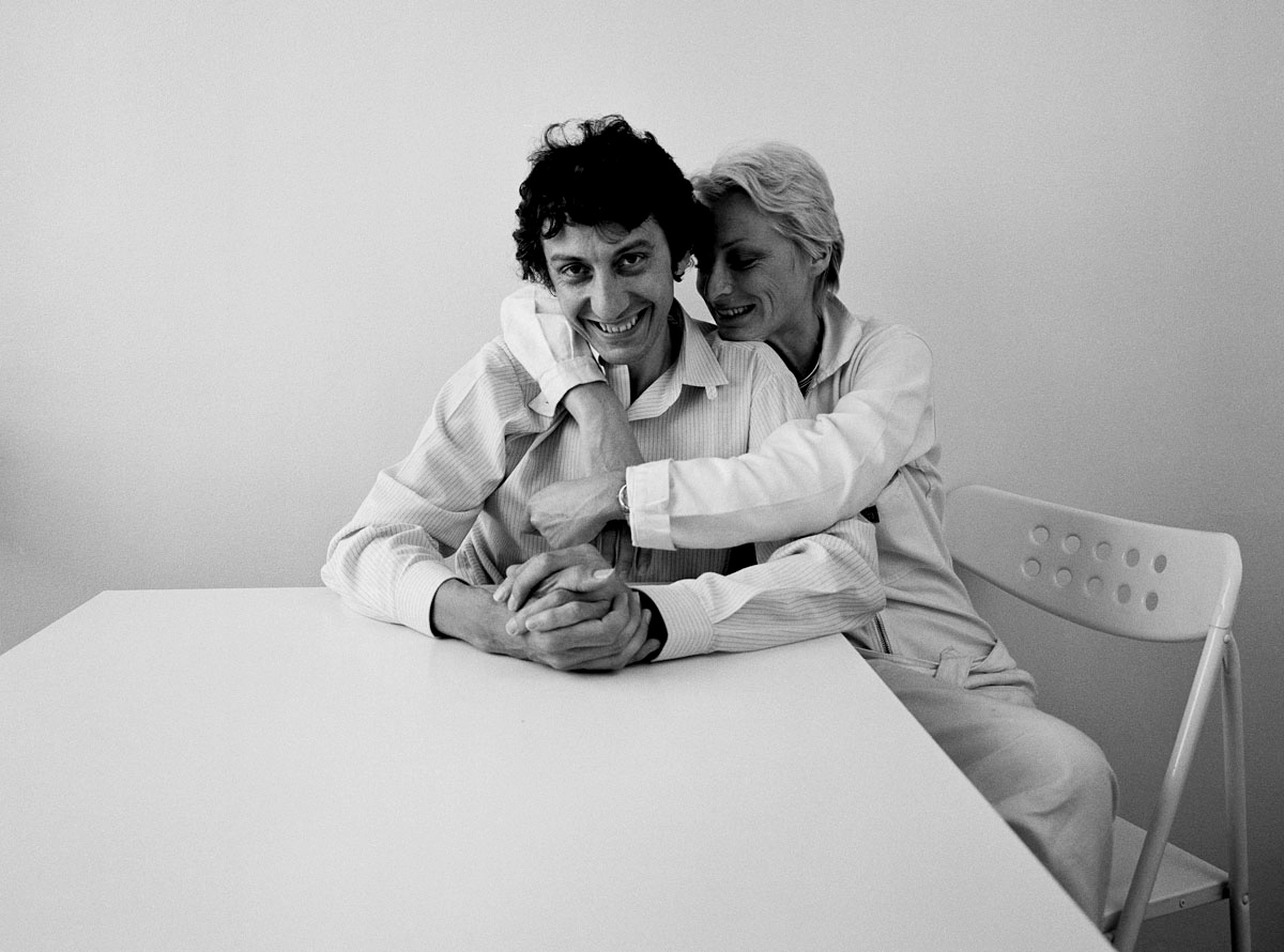 La photographie montre un couple enlacé vêtus de blanc assis à une table.
