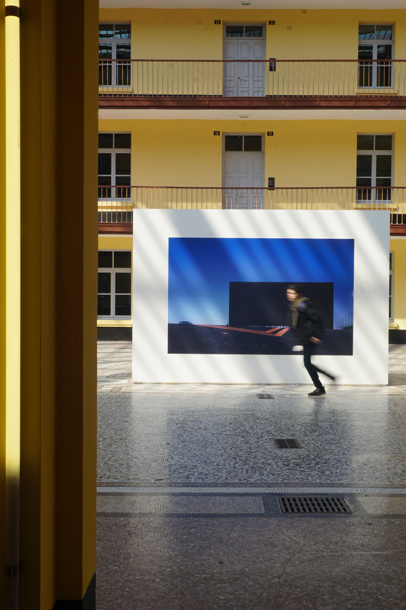 La photographie montre une cimaise installée dans une cour ensoleillée